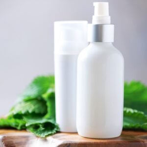 Nettle lotion, cream, shampoo or soap in white bottle and fresh nettles leaves on grey background