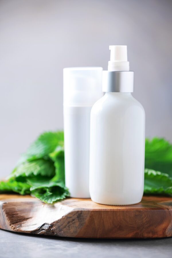 Nettle lotion, cream, shampoo or soap in white bottle and fresh nettles leaves on grey background